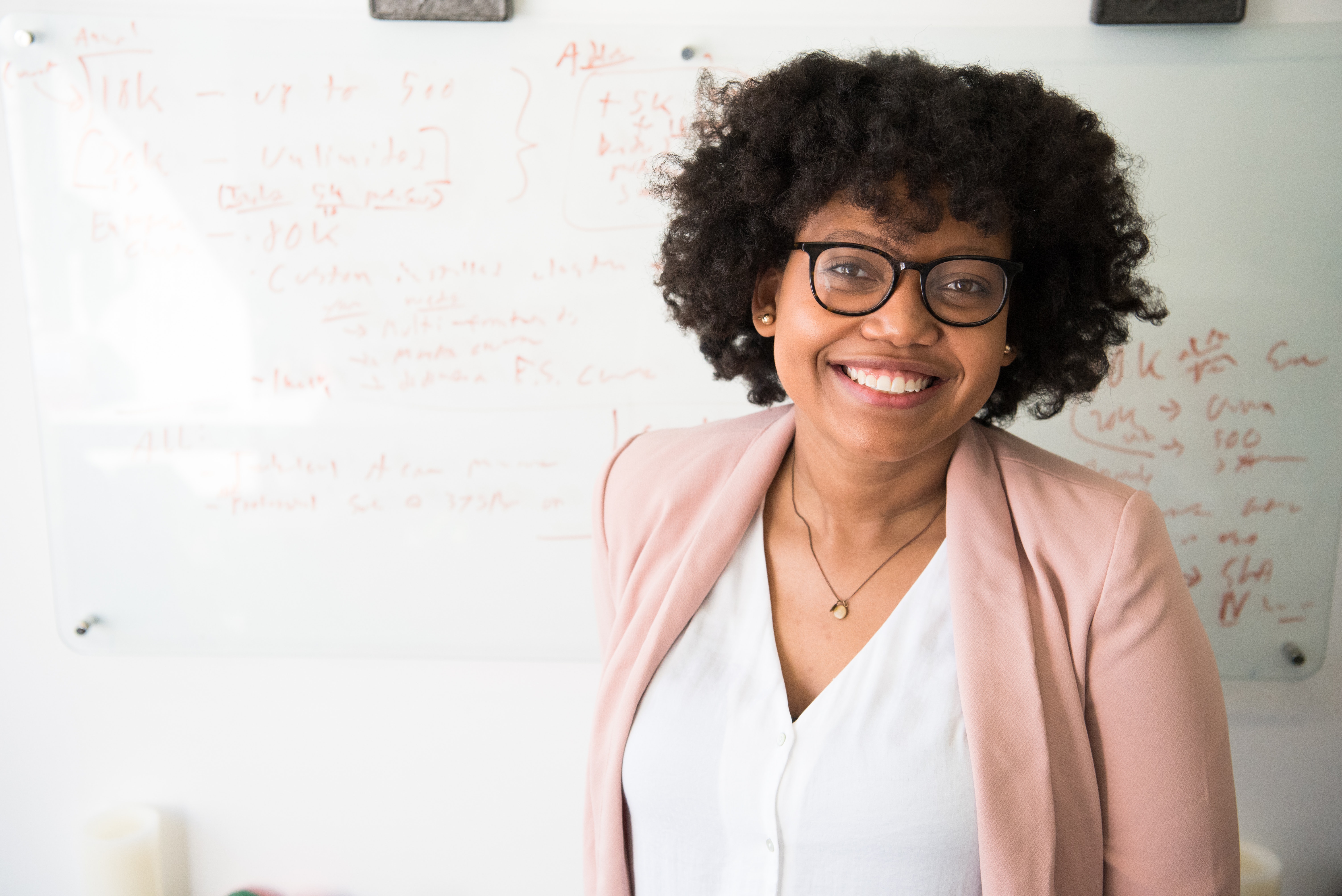 Woman in glasses, business attire and a pink jacket smiling at the camera.