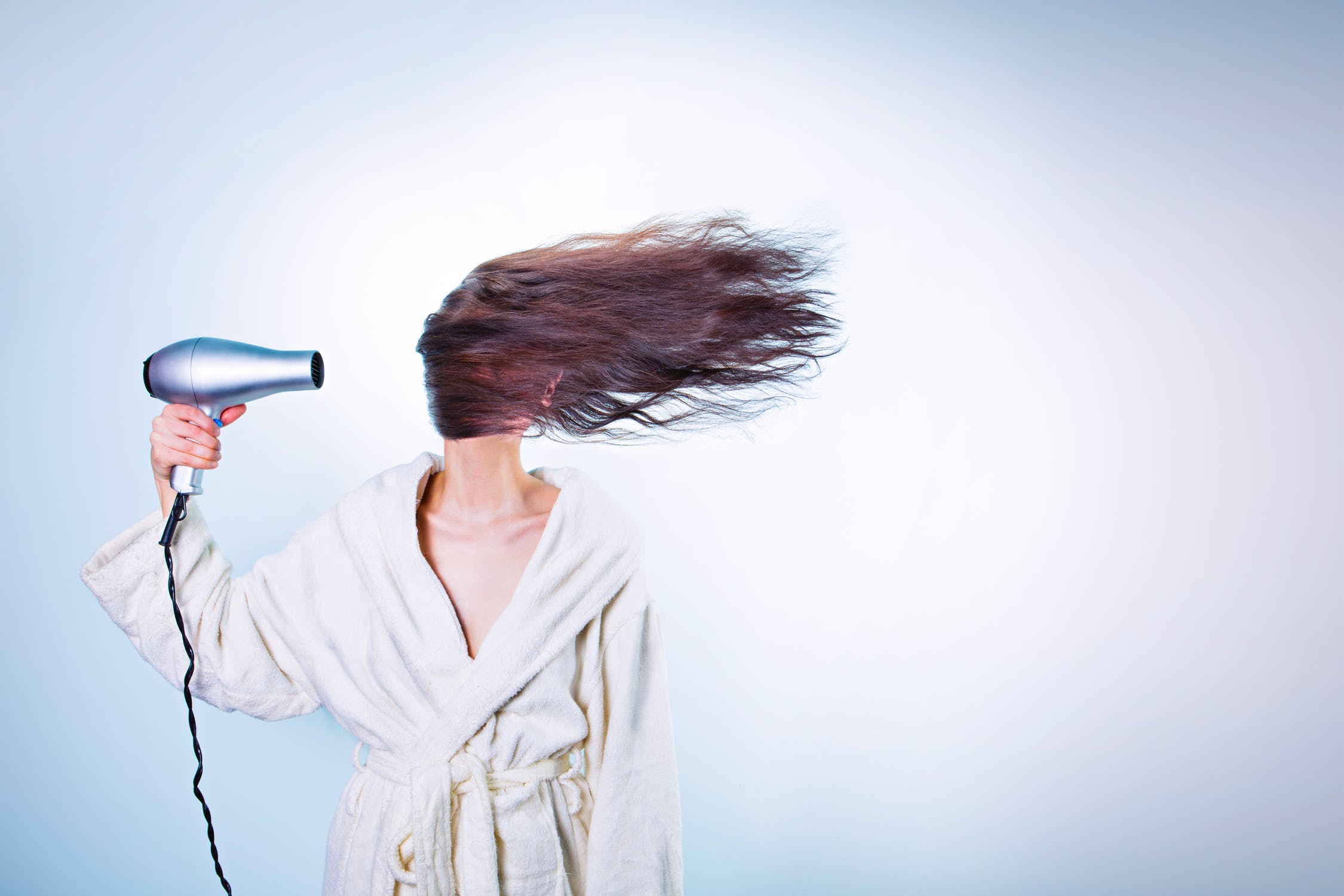 Woman in a robe with a hair dryer pointed at her head, blowing her hair to the side.